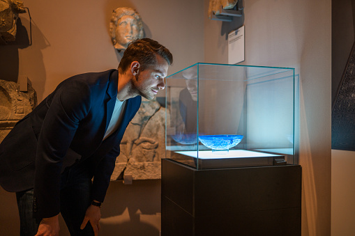 Attractive Caucasian man leaning forward to get a better look at the blue bowl exhibited in a glass case. Waist up image, his face lit up by the display lights, looking away.