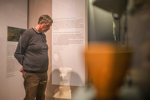 A senior Caucasian male museum visitor admiring the objects in glass cases. 3/4 length image, shot through glass, side view of the man.