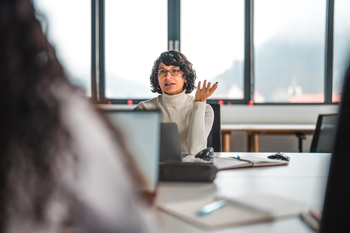 A portrait of a mid adult Latina business developer  at one side of an office desk, motivating a long-haired female employee across the table. Waist up image, looking away. Open laptops  in front of women.