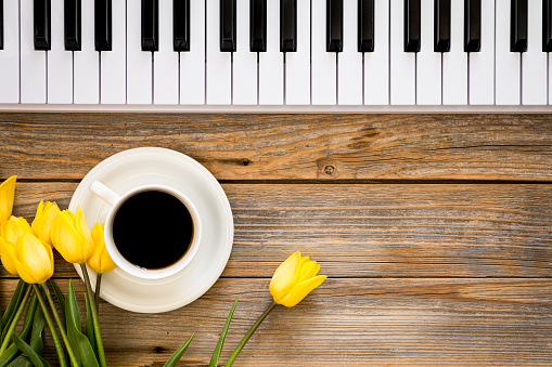 Musical background with white piano keys, a cup of black coffee and a bouquet of yellow tulip flowers on a wooden surface, top view, copy space.