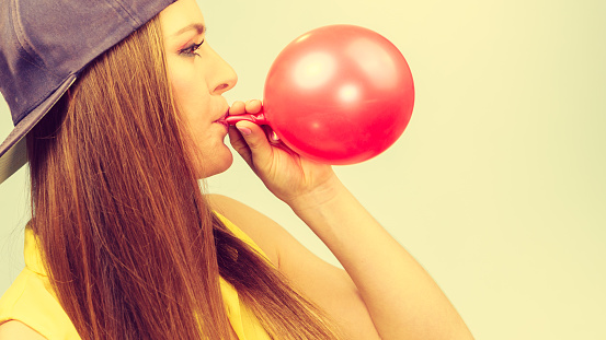 Time for party. Young trendy woman prepare birthday celebration decoration. Teenage girl in cap blowing red balloon.