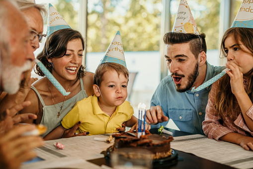 A small boy celebrating a birthday and blowing a candle surrounded by his big family.