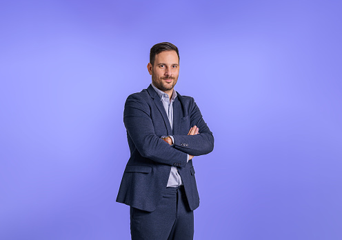 Portrait of handsome male professional executive wearing elegant blue suit and posing confidently. Smiling businessman with arms crossed looking at camera and standing against blue background