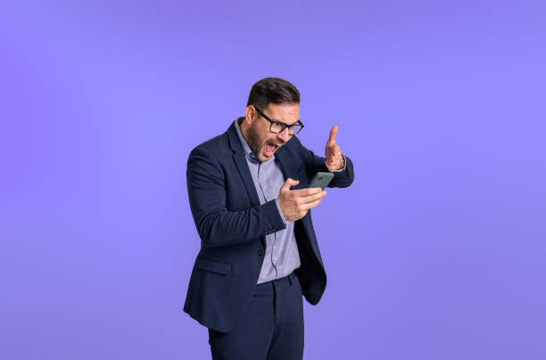 istock Devastated male professional manager dressed in suit shouting angrily and pointing at mobile phone. Irritated despair young businessman yelling at cellphone isolated against blue background 1491879896