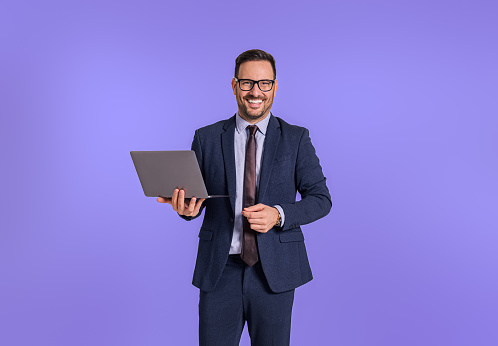 Portrait of smiling male professional manager dressed in formalwear holding laptop and looking at camera. Confident businessman working over wireless computer while standing against blue background
