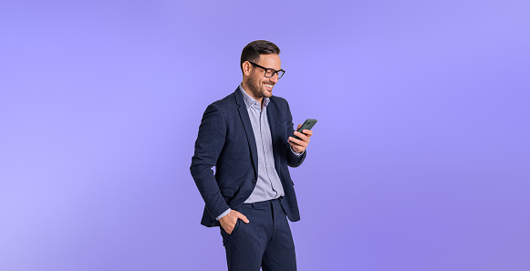 Male business professional dressed in elegant formals smiling and messaging online over mobile phone. Handsome chief with hand in pocket using cellphone while posing against blue background