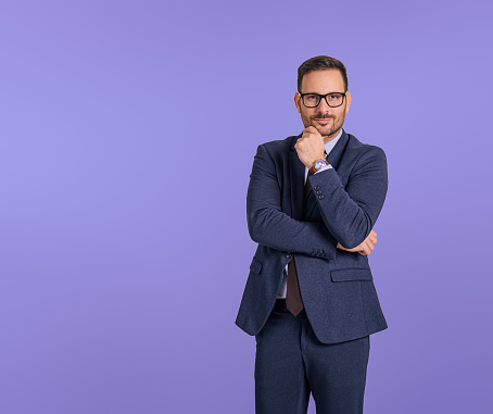 Handsome young professional manager touching chin and folding hand while posing against blue background. Portrait of male entrepreneur dressed in elegant formals looking at camera confidently