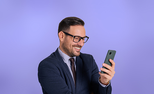 Portrait of smiling young business executive text messaging over smart phone. Charming happy male manager dressed in elegant formals chatting over cellphone against isolated blue background