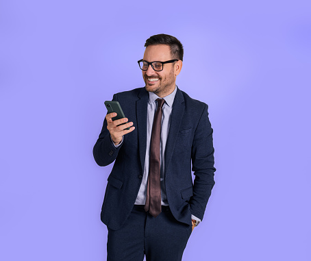 Charming male business professional with hand in pocket checking messages over smart phone. Young entrepreneur dressed in formals scrolling social media on cellphone isolated against blue background