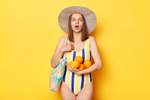 Shocked woman wearing swimsuit isolated on yellow background holding oranges being surprised of high price for fruit looking at camera with open mouth.