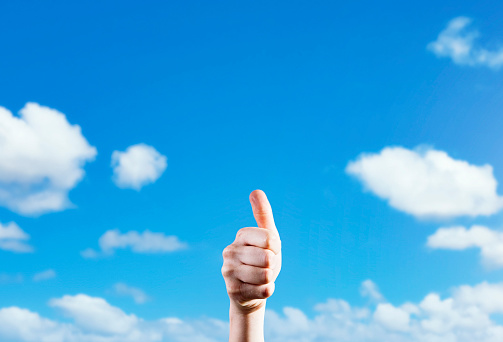 Thumbs-up sign into clear sky against scattered summer clouds.