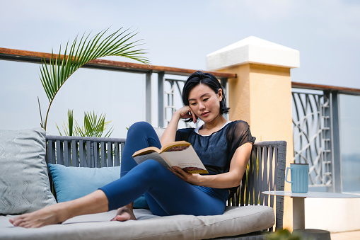 Young Asian woman sitting comfortably on a lounge chair and reading a book on the balcony, feeling relaxed. Enjoying her quiet time at home