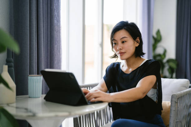 istock Young Asian woman sitting at table by the window and using digital tablet while working from home. Home office 1491890789