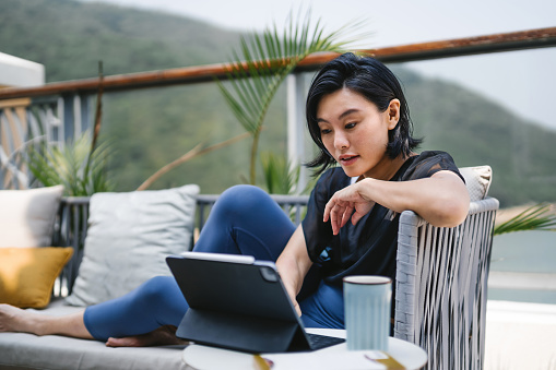 Young Asian woman sitting on the balcony, using digital tablet and having a cup of coffee in the morning. Young female using computer for leisure or business at home