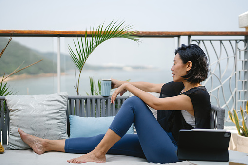 Young Asian woman sitting comfortably on a lounge chair with cup of coffee, enjoying a relaxing moment looking at the sea and morning sunlight on her balcony