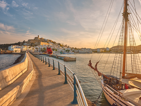 Wide-angle view of Eivissa’s harbour, old town and marina at sunset. Picturesque clouds, calm waters reflecting the warm light of a summer evening, a moored sailing ship and fast ferries, a long jetty, the iconic skyline of Dalt Vila dominated by the cathedral church of Santa Maria de les Neus. High level of detail, natural rendition, realistic feel. Developed from RAW.