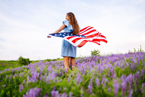 Young woman waves an american flag on blooming meadow.