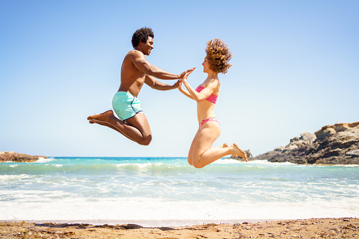 Full body side view of cheerful African American boyfriend and curly haired girlfriend, in swimwear jumping on sandy beach and holding hands near waving sea while looking at each other