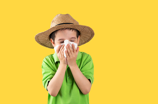 Child in hat blowing into wipe suffering from flu symptoms on yellow background.