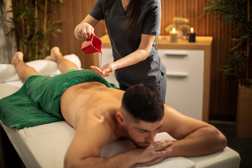 Mid adult man relaxing in spa center and receiving back and shoulder massage. Massage therapist melting wax from candle and applying it warm on skin. Nice cozy space with candles in background and professional therapist make him feeling wonderful