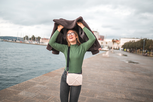 3/4 length image of a mid adult Caucasian woman with glasses smiling while keeping  her hair away from the rain by covering her head with a jacket. Sea and seaside town in the background.