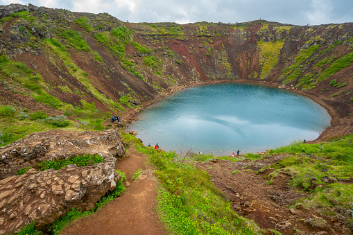 Top view of Kerid Crater Lake in Iceland on a cloudy day with people walking and standing around in the distance. Turquoise waters of Icelandic crater lagoon