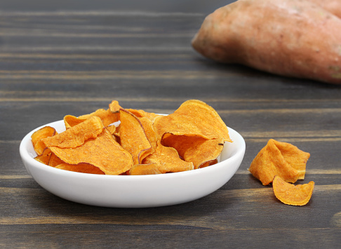 A bowl of sweet potato chips on a wooden table with a sweet potato in background.  Macro with copy space.