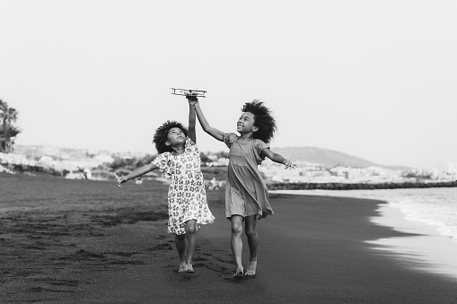 Afro twins sisters running on the beach while playing with wood toy airplane - Focus on faces - Black and white editing