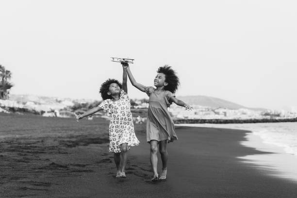 irmãs gêmeas afro correndo na praia enquanto brincam com avião de brinquedo de madeira - foco nos rostos - edição em preto e branco - airplane black and white fun child - fotografias e filmes do acervo