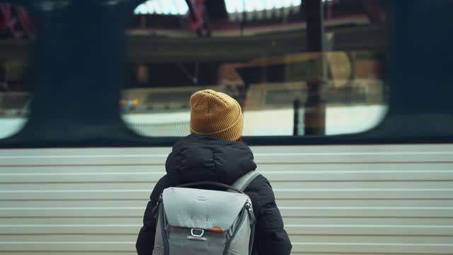 Rear view of young woman with backpack while waiting at platform of train station looking at passing tram, slow motion.