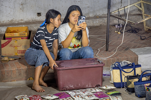 Night Market, Sisavangvong Road, Luang Prabang, Laos - March 21th 2023: Two young women studying the content of a mobile phone while waiting for customers in their shop at the famous night market which primary is selling clothes to the tourists