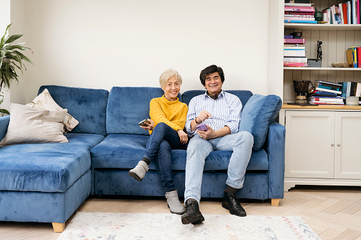 Contented and happy Asian couple in their 60s and 70s relaxing at home sitting of sofa in their living room smiling.
