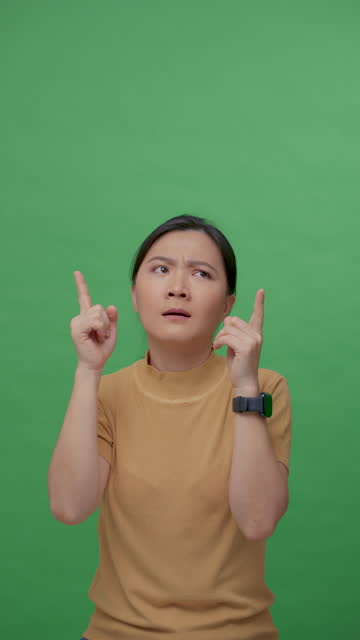 Asian woman feel confused, thinking and pointing finger up standing isolated over green background.