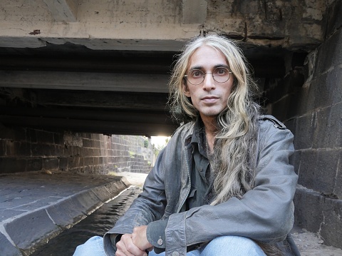 Man with long hair in leather jacket posing under antique bridge