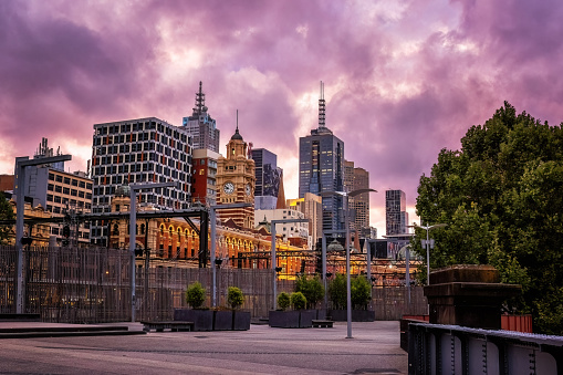 Melbourne cityscape, with Flinders Street train station in the foreground, at dusk from the walkway along the banks of the Yarra river. Bright pink sunset light and sky.