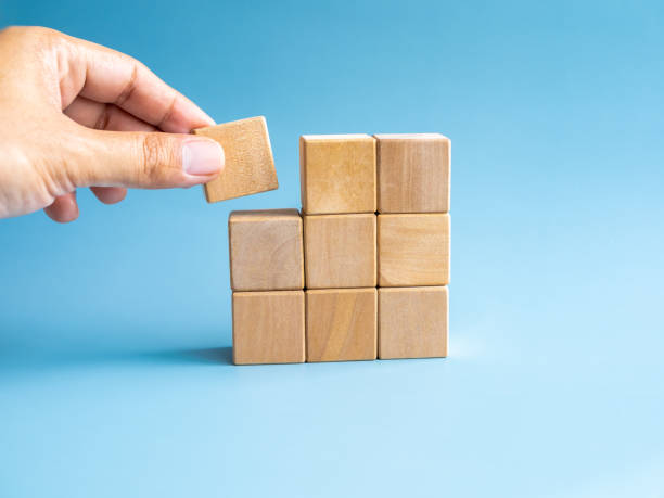 istock Last piece wood cube block in hand putting on wooden puzzles stacking arranging compleated isolated on blue background, minimal style. Solution, solve problem, business goal and success plan concepts. 1492022311