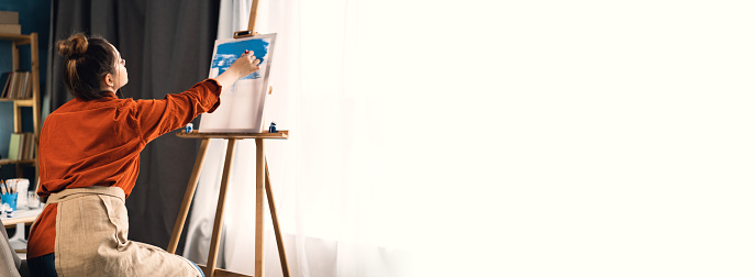 young brazilian woman artist in home studio sitting on a chair drawing a picture. Black hair, dressed in casual clothes. concept of creativity and hobby. Banner. copy space.
