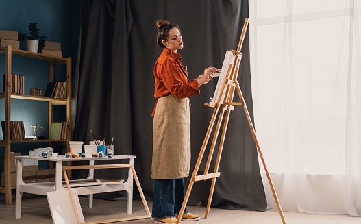 Beautiful female painter with easel in art studio. Young talented artist working. Copy space