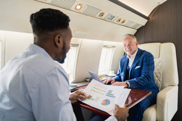 Black businessman in early 30s holding printout of graphs and conversing with Caucasian associate in mid 50s as they fly in comfort.