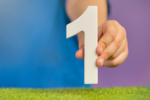 Number 1 in hand. Hand holding white number one over green grass and purple background, birthday concept, first or winner, first of January, Canada Day, copy space. High quality photo