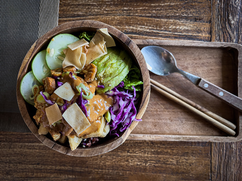 Vegan tempeh rice and vegetable bowl topped with wonton wrappers and peanut sauce in a wood bowl and tray.  Bali, Indonesia.