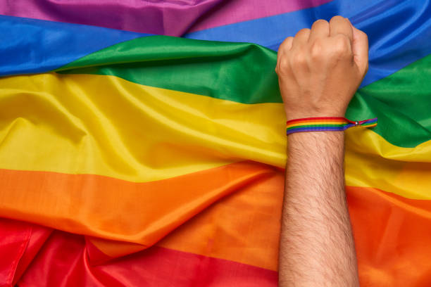 pugno dell'attivista con braccialetto lgbt sullo sfondo della bandiera arcobaleno - gay pride wristband rainbow lgbt foto e immagini stock