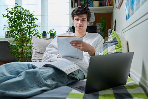 Teenage guy lying at home on couch student using laptop notebook textbook. Handsome male teenager college, high school student learning online remotely. Education, learning, technology, youth concept