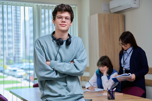 Portrait of college student guy looking at camera inside classroom. Young male 18, 19 years old wearing glasses, with headphones, posing in education center. Education, knowledge, learning, youth concept