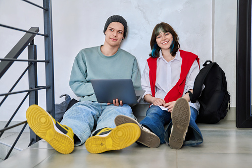 Friends hipster teenagers guy and girl sitting on floor with backpacks using laptop looking at camera inside study hall corridor. Fashion trendy youth, lifestyle, communication, friendship concept