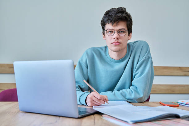 istock Young guy college student sitting at desk in classroom, using laptop, looking at camera 1492051613