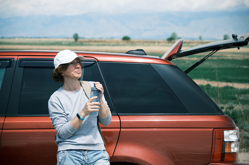 Woman is drinking water next to the car standing in the fields