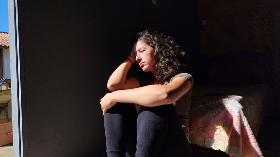 Depressed and anxious woman, sitting on the floor leaning against the edge of the bed, sunlight coming through the door. Made in Maio, Jaguaruna, SC, Brazil.