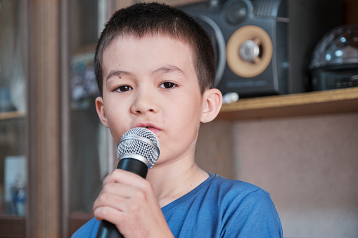 Close-up portrait of serious Asian boy looking at camera holding microphone singing song karaoke at home concert. Children's lifestyle concept