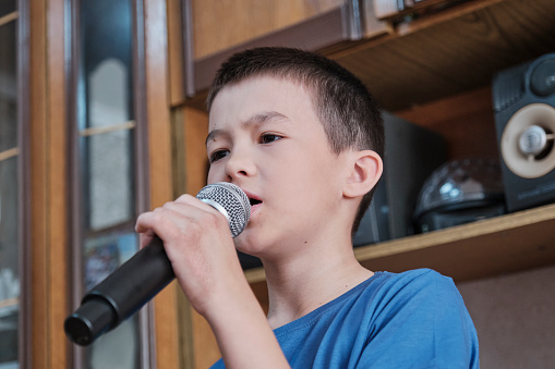Close-up portrait of serious Asian boy holding microphone singing song karaoke at home concert. Children's lifestyle concept